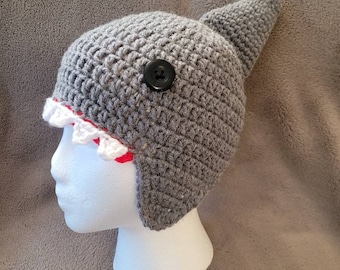 May include: Gray crocheted shark hat with a red and white trim and a black button. The hat is on a white mannequin head.
