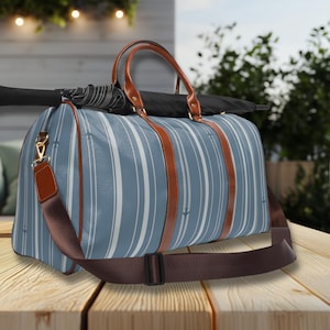 Puede incluir: Una bolsa de lona a rayas azules y blancas con asas y detalles de cuero marrón. La bolsa tiene una correa de hombro marrón desmontable y herrajes dorados. Una sombrilla negra descansa sobre la bolsa. La bolsa está sobre una superficie de madera.