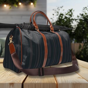 Puede incluir: Bolsa de viaje a rayas negras y grises con detalles en cuero marrón. La bolsa tiene una correa para el hombro y asas marrones. Un paraguas descansa encima. La bolsa está sobre una superficie de madera.
