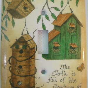 May include: A light switch cover with a beige background and a whimsical design of three birdhouses. The birdhouses are painted in shades of brown and green. The switch cover features the text "The Earth is full of the Goodness of the Lord Psalm 33:5".
