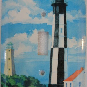 May include: A light switch cover featuring a watercolor painting of two lighthouses against a blue sky with white clouds. The larger lighthouse is black and white striped and the smaller lighthouse is yellow. There are green trees and a white picket fence in the foreground.