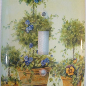 May include: A light switch cover with a painted design of three potted topiary trees with blue flowers. The background is a light green color.