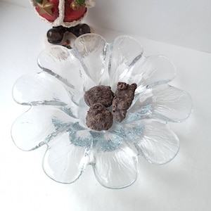 May include: A clear glass flower-shaped bowl contains three pieces of chocolate. A snowman figurine in a red coat, green scarf, and a red hat stands in the background. The snowman is holding a gold star.