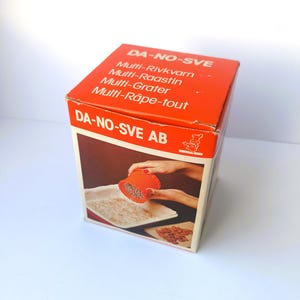 May include: A vintage orange and white box for a multi-grater, with text in multiple languages. The box features an image of the grater in use, with a person grating food into a tray. The brand name "DA-NO-SVE AB" is also visible.