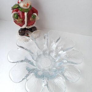 May include: A clear glass flower-shaped bowl with textured petals. A snowman figurine in a red coat, green scarf, and a red hat stands behind the bowl. The snowman holds a gold star and a green ball.