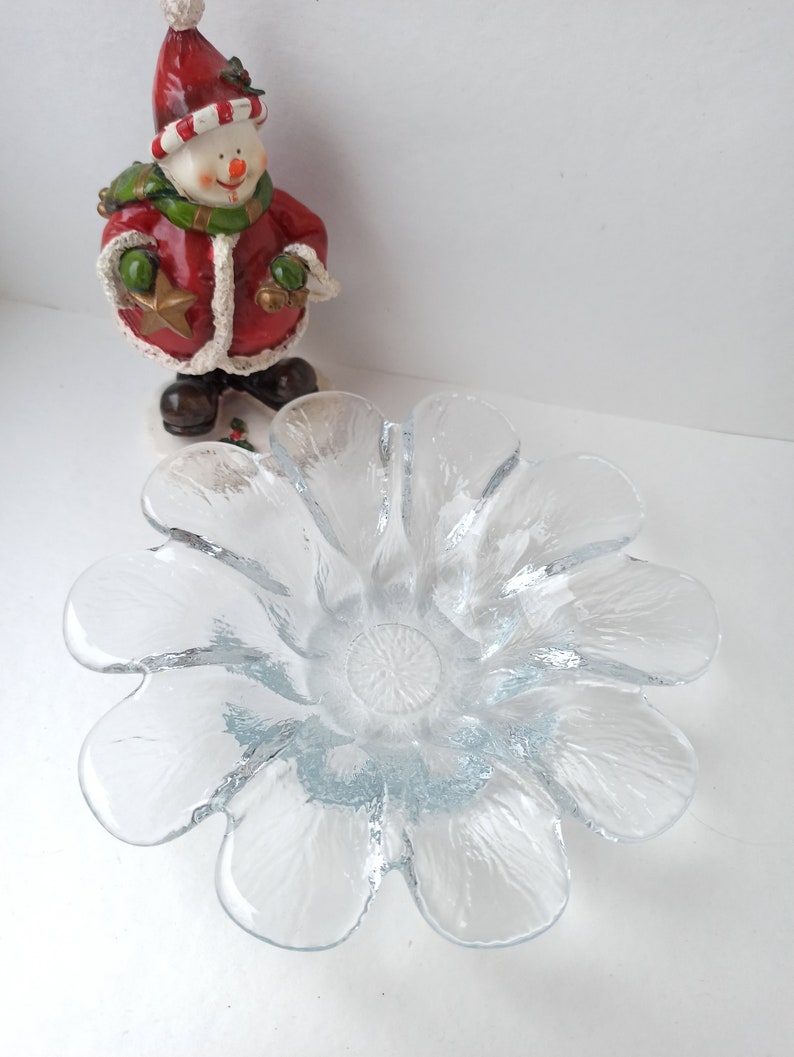 May include: A clear glass flower-shaped bowl with textured petals. A snowman figurine in a red coat, green scarf, and a red hat stands behind the bowl. The snowman holds a gold star and a green ball.