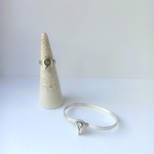 May include: A silver ring and a matching silver bracelet, both featuring a teardrop-shaped centerpiece with a small embedded clear stone. The ring is displayed on a speckled cream-colored cone, while the bracelet lies flat.