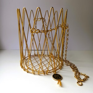 Può includere: Un cestino in filo metallico color oro di forma cilindrica con un motivo incrociato. Il cestino presenta una base circolare con un design a griglia ed è sospeso da una catena in metallo color oro. È attaccata una piccola etichetta rotonda con il testo ARTICLE GARANTI OR FIN.