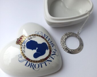 May include: A white, heart-shaped trinket box with a matching lid. The box features a blue silhouette design with gold accents and text. A silver-toned, circular charm with a chain is next to the box.