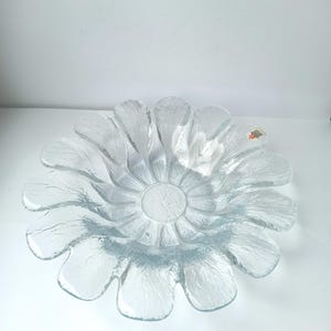 May include: Clear glass flower-shaped bowl with textured petals. The bowl has a central circular depression and a textured surface. The petals are evenly spaced around the centre, creating a floral design. The bowl is likely for decorative or serving purposes.