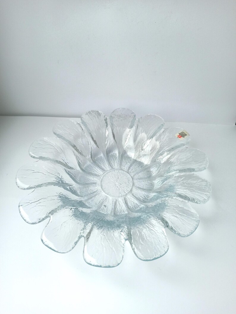 May include: Clear glass flower-shaped bowl with textured petals. The bowl has a central circular depression and a textured surface. The petals are evenly spaced around the centre, creating a floral design. The bowl is likely for decorative or serving purposes.