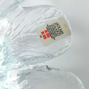 May include: A clear glass decorative object with a petal-like design. The object has a sticker that reads "HOLME GAARD COPENHAGEN" and "MADE IN DENMARK". The glass has a textured surface and is partially transparent.