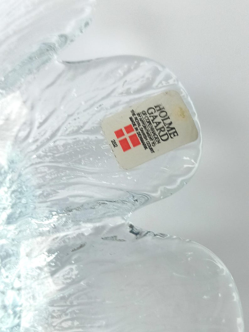 May include: A clear glass decorative object with a petal-like design. The object has a sticker that reads "HOLME GAARD COPENHAGEN" and "MADE IN DENMARK". The glass has a textured surface and is partially transparent.
