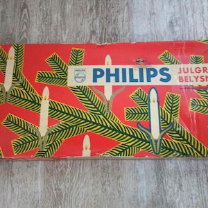 May include: Vintage Philips Christmas light box. The red box features a pattern of green fir branches and white candles. The Philips logo is visible, along with the text "JULGRANS BELYSNING". The box is rectangular and shows signs of wear.