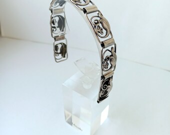 Könnte beinhalten: Ein silbernes Armband mit einem zarten, filigranen Design mit sich wiederholenden floralen Motiven. Das Armband ist offen und hat einen Scharnierverschluss.