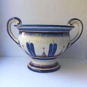 Könnte beinhalten: Eine Keramikvase mit zwei geschwungenen Griffen, mit einer weißen Basis mit blauen und orangefarbenen floralen Designs. Der Rand und die Griffe sind mit Blau akzentuiert, und die Vase steht auf einem Sockel. Ein dekoratives Stück für die Inneneinrichtung.