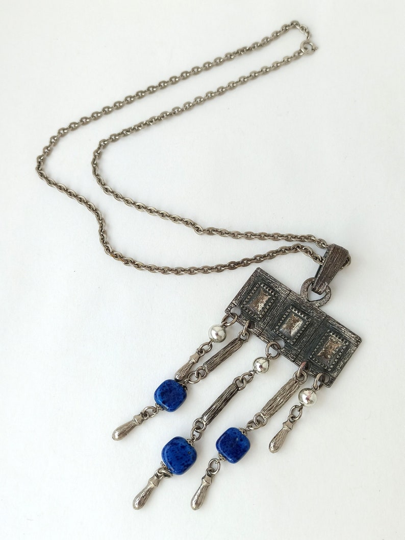 May include: A silver chain necklace with a rectangular pendant featuring a geometric design and blue glass beads hanging from it.