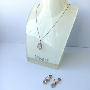 Peut inclure: Un collier doré avec un pendentif ovale bleu clair est présenté sur un buste blanc. Des boucles d'oreilles assorties à clip avec des pierres bleues similaires sont en dessous. Le mot "Misaki" est imprimé sur le buste.