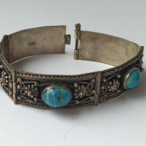 May include: Silver-toned hinged bracelet with three oval turquoise-colored stones. The bracelet features intricate filigree detailing and a secure clasp. The design is vintage-inspired, with a classic and elegant aesthetic.