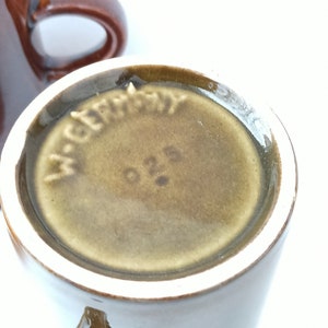 Puede incluir: Primer plano de la parte inferior de una taza de cer&aacute;mica marr&oacute;n con la palabra "Alemania" y el n&uacute;mero "026" impresos en la parte inferior.