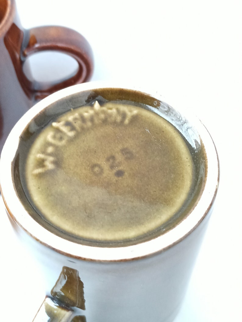 Puede incluir: Primer plano de la parte inferior de una taza de cer&aacute;mica marr&oacute;n con la palabra "Alemania" y el n&uacute;mero "026" impresos en la parte inferior.