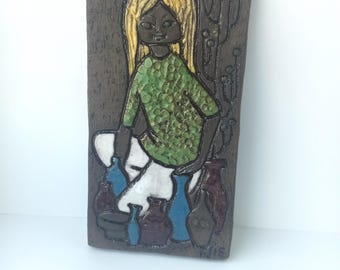 May include: A ceramic wall plaque featuring a girl with blonde hair and a green shirt, surrounded by colorful pottery. The plaque is brown and has a leather strap for hanging.