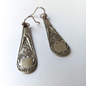 Peut inclure: Une paire de boucles d'oreilles pendantes argentées. Chaque boucle d'oreille a une forme de goutte d'eau avec des détails floraux et des volutes ornés. Les boucles d'oreilles ont une fermeture à crochet et un espace rectangulaire en bas.