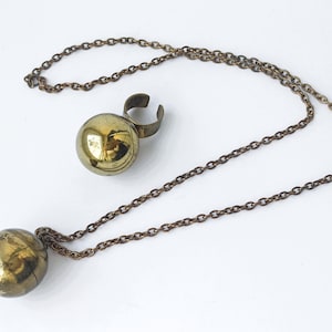 May include: A vintage jewelry set featuring a necklace and a ring. The necklace has a chain with a round, gold-colored pendant. The ring has a matching gold-colored ball. The metal has a tarnished, antique appearance.