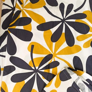 Peut inclure: Un tissu textile présentant un motif répétitif de grandes formes stylisées ressemblant à des fleurs, en jaune moutarde et gris anthracite foncé sur un fond blanc. Le design se compose de multiples pétales arrondis partant d'un centre.