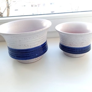 以下が含まれることがあります： 中央に青い帯が巻かれた白い陶器製のプランターが2つ。左側に大きいプランター、右側に小さいプランターがあります。