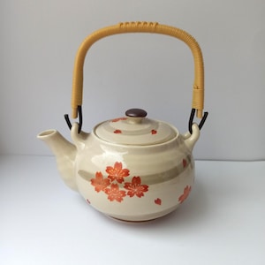 Op de afbeelding: Crèmekleurige keramische theepot met een gevlochten handvat en deksel. De theepot is versierd met rode kersenbloesemmotieven en bruine accenten. De handgreep is gemaakt van gevlochten stro.