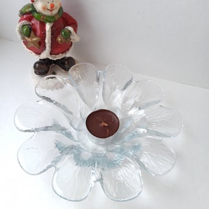 May include: A clear glass flower-shaped candle holder with a small brown candle in the centre. A snowman figurine in a red coat and hat stands in the background. The candle holder is a decorative item.