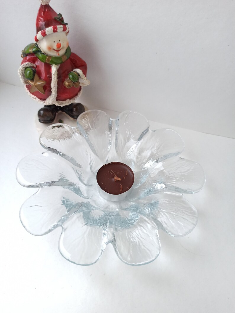 May include: A clear glass flower-shaped candle holder with a small brown candle in the centre. A snowman figurine in a red coat and hat stands in the background. The candle holder is a decorative item.