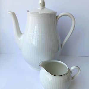 May include: A white ceramic tea set with a textured, vertical striped pattern. The set includes a tall teapot with a lid and a small creamer. Both pieces have silver trim around the edges.