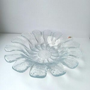 May include: Clear glass bowl shaped like a flower. The bowl has a textured surface and petal-like edges. The center of the bowl has a circular, textured design. The bowl is on a white surface.