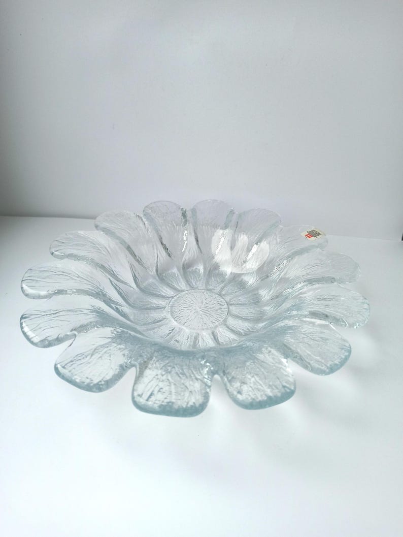 May include: Clear glass bowl shaped like a flower. The bowl has a textured surface and petal-like edges. The center of the bowl has a circular, textured design. The bowl is on a white surface.