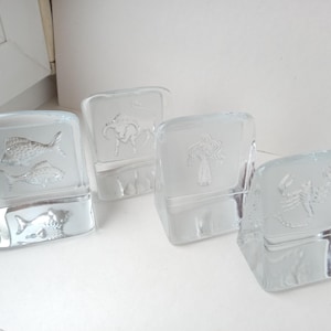 May include: Four clear glass blocks with etched designs. One block features two fish, another a bull, a third a tree, and the last a scorpion. The blocks are arranged on a white surface, showcasing the detailed artwork.