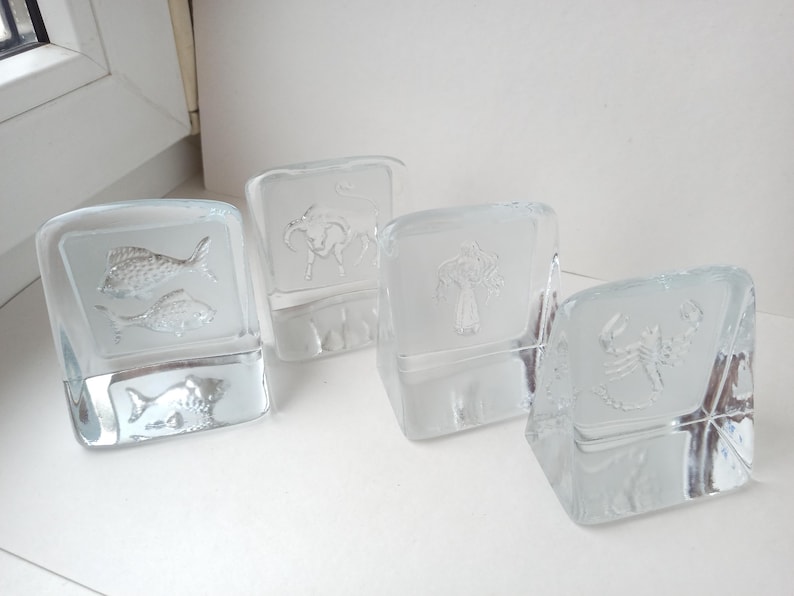 May include: Four clear glass blocks with etched designs. One block features two fish, another a bull, a third a tree, and the last a scorpion. The blocks are arranged on a white surface, showcasing the detailed artwork.