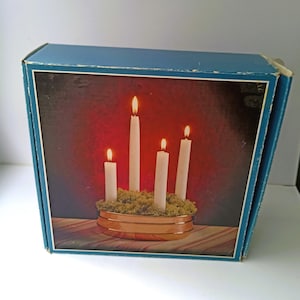 May include: A blue cardboard box featuring a printed image of four white taper candles of varying heights, each with a lit flame, arranged in a copper-colored circular bowl filled with green moss against a dark red background.
