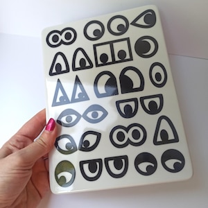 May include: A white rectangular tray with a glossy finish, featuring a collection of black eye illustrations in various shapes and sizes. The eyes include circles, triangles, and rectangles. The tray is held by a person.