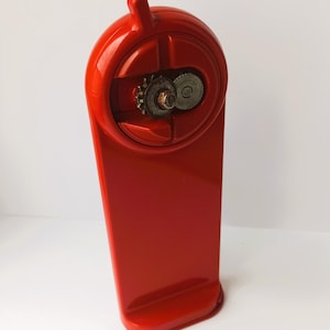 May include: A bright red, vintage-style can opener. The opener has a tall, rectangular body with a rounded top section housing the cutting mechanism. The gears and cutting wheel are visible. The can opener is designed to be mounted on a counter.