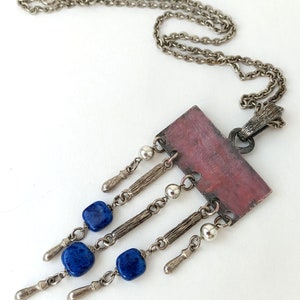 May include: A silver chain necklace with a rectangular pendant featuring a pink and brown design. The pendant has five silver chains with blue gemstone beads and silver accents hanging from it.