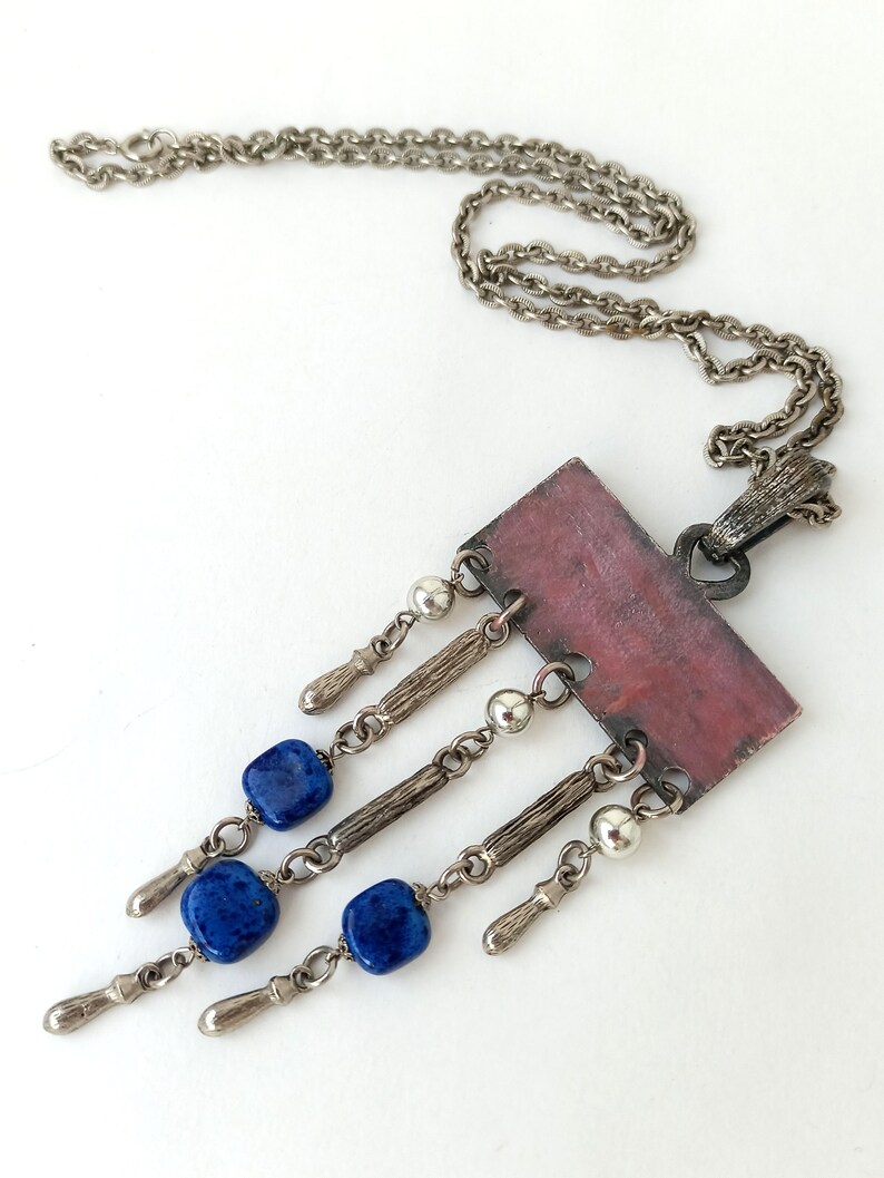 May include: A silver chain necklace with a rectangular pendant featuring a pink and brown design. The pendant has five silver chains with blue gemstone beads and silver accents hanging from it.