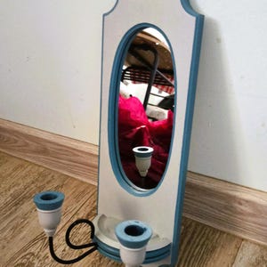 May include: A wall-mounted candle holder featuring a vertical white and blue rectangular frame with an oval mirror in the center. Two black metal arms extend from the base, each supporting a white and blue candle cup for holding taper candles.