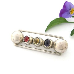 Peut inclure: Une broche en argent avec cinq pierres rondes disposées en ligne. Les pierres sont rouges, jaunes, violettes et deux argentées.