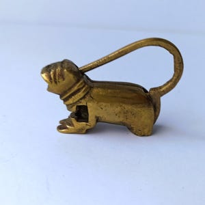 May include: A small, brass-colored metal figurine of an animal, possibly a rodent, with a loop extending from its back. The figurine has detailed features and is set against a white background. The loop suggests it may be a keychain or decorative item.