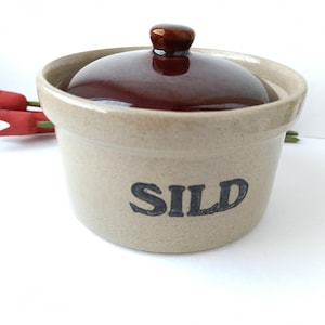 May include: A beige ceramic crock with a brown lid and the word "SILD" written in black lettering. The crock is sitting on a white surface.