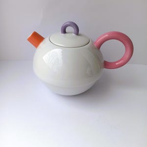 May include: A white ceramic teapot with a round body, orange spout, pink handle, and purple lid handle. The teapot has a glossy finish and a modern design, suitable for serving tea.