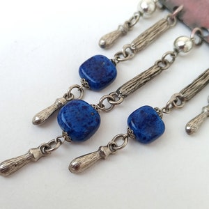 May include: A silver and blue statement necklace with a rectangular pendant and multiple dangling charms. The charms include small silver beads, silver teardrop shapes, and square blue glass beads with a speckled pattern.