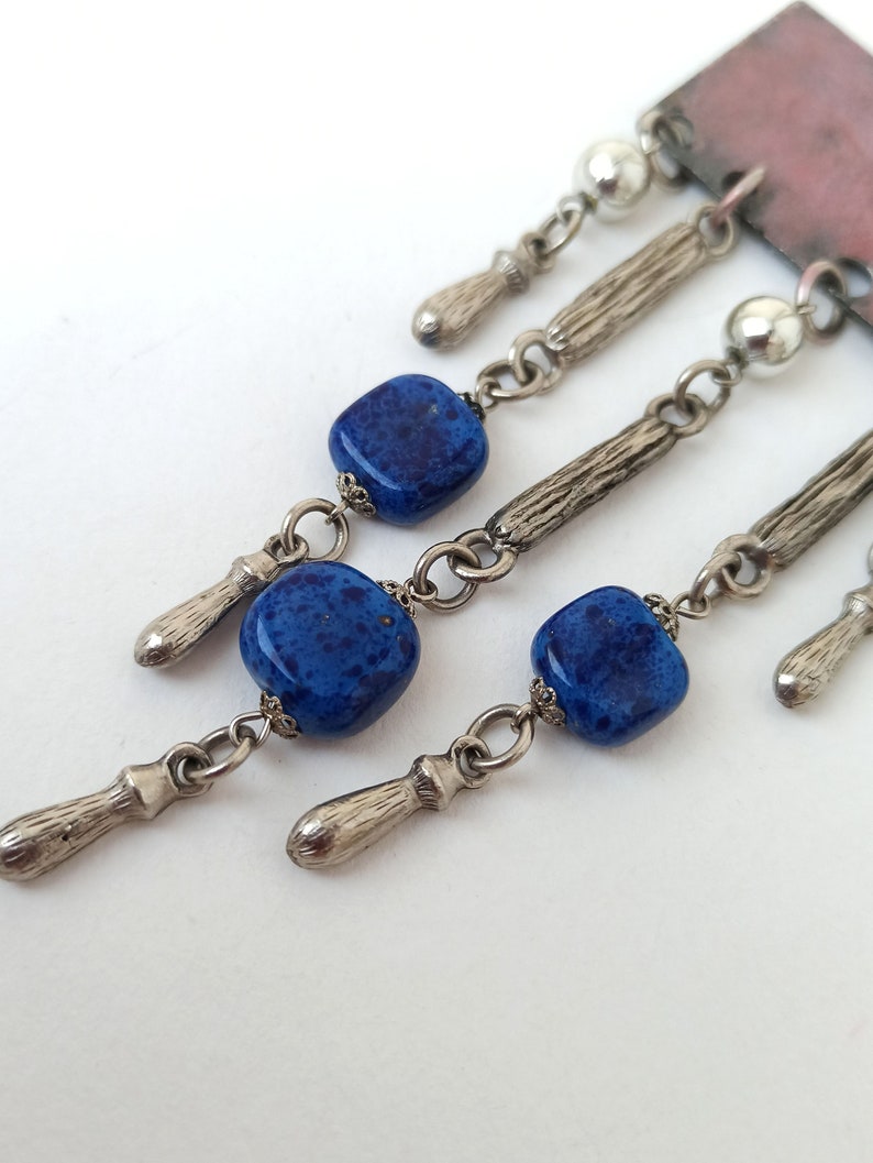 May include: A silver and blue statement necklace with a rectangular pendant and multiple dangling charms. The charms include small silver beads, silver teardrop shapes, and square blue glass beads with a speckled pattern.
